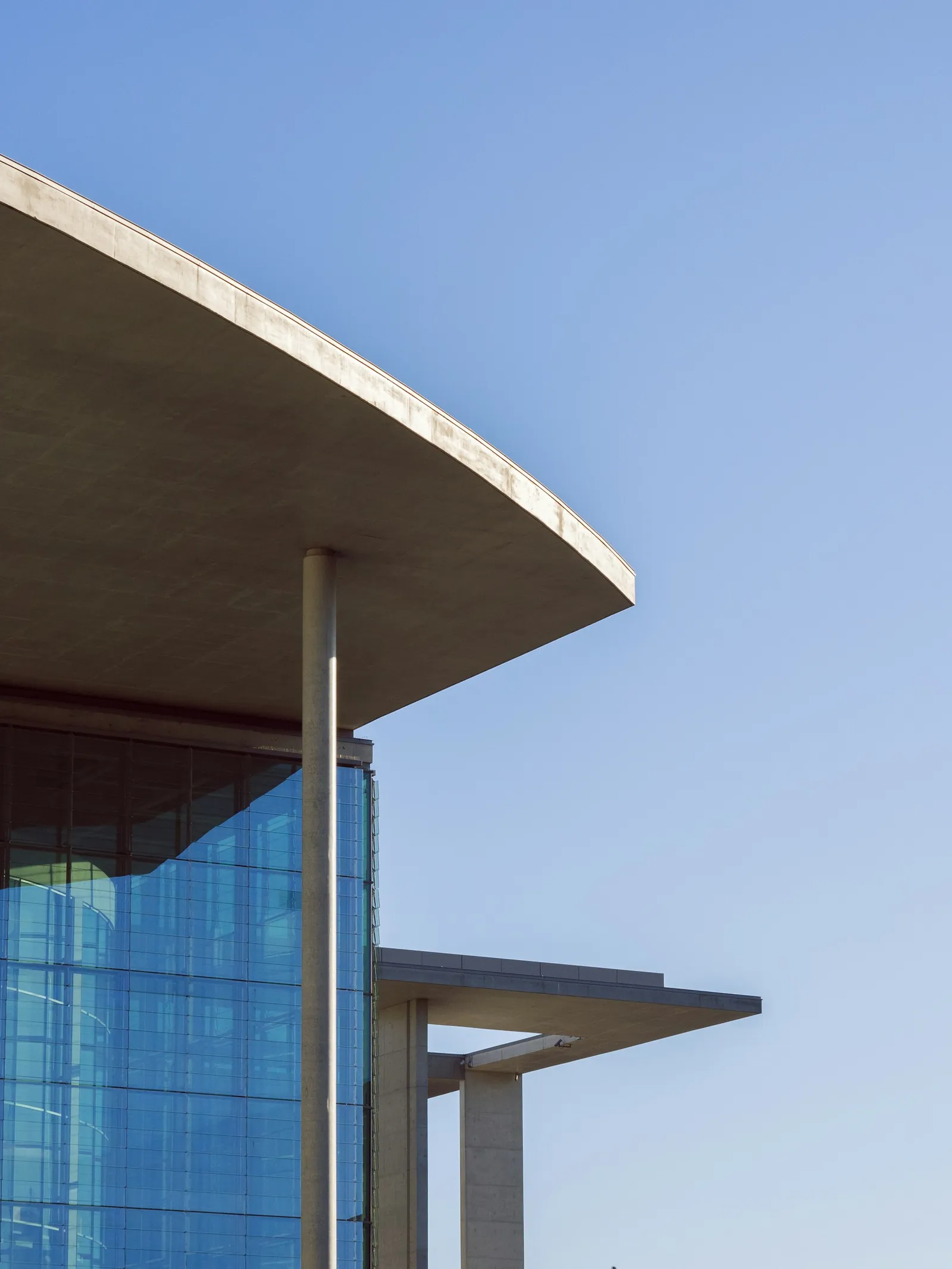 Auskragendes Betondach eines modernen Gebäudes mit blau spiegelnder Glasfassade und schlanker Stütze vor klarem Himmel